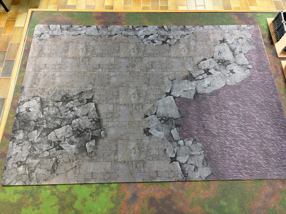 A rectangular tabletop gaming terrain mat featuring a printed scene of gray stone ruins breaking into a purple grassy field, displayed on a green and brown camouflage-patterned gaming mat over a tiled floor.